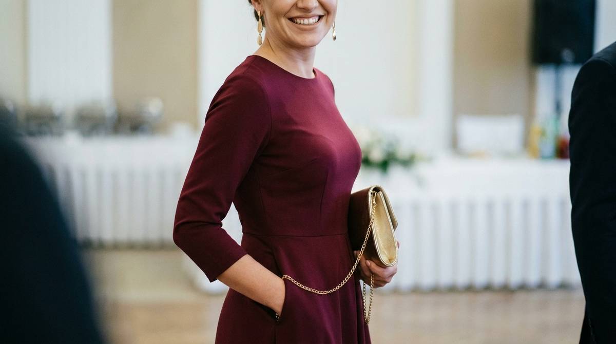 Kobieta w bordowej sukience trzymająca złotą torebkę na wesele